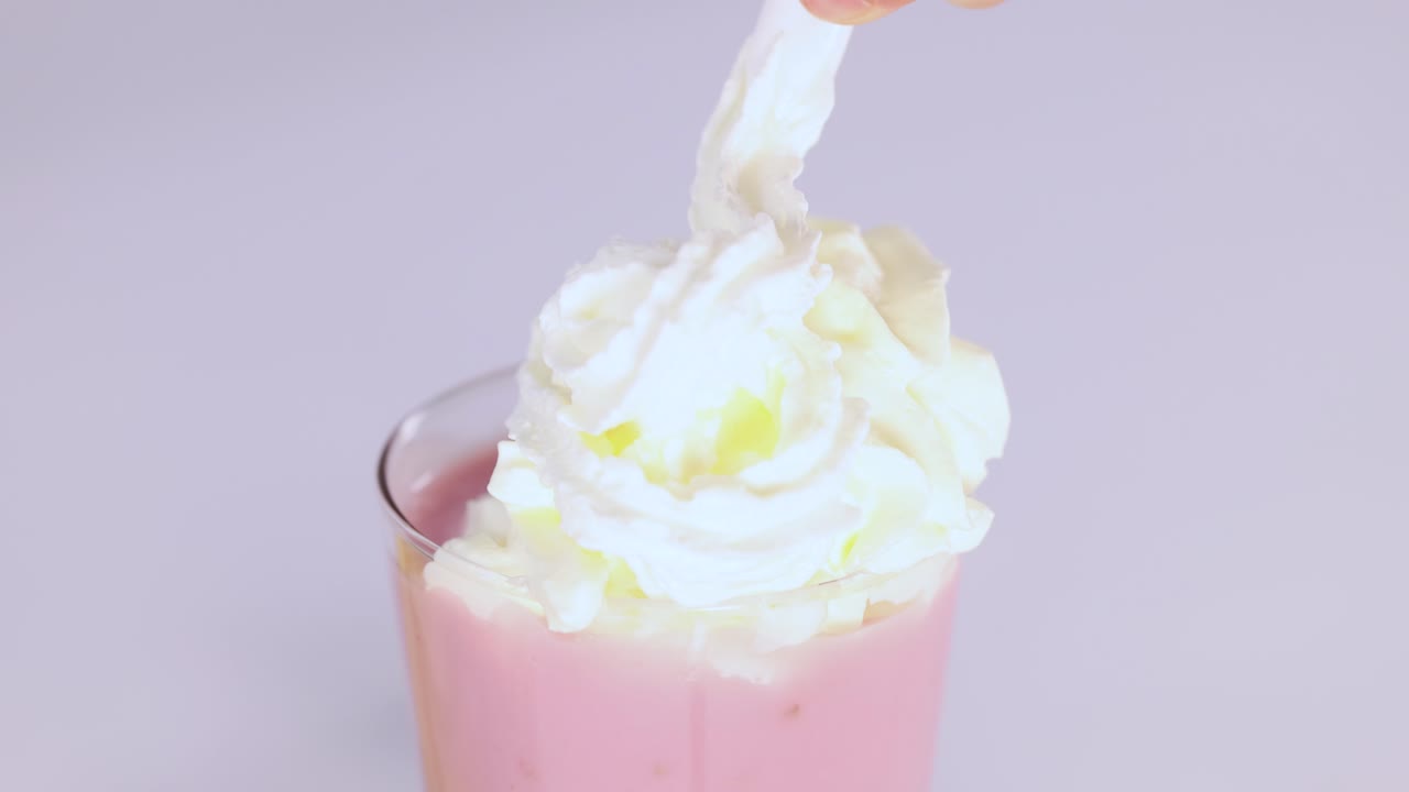Una mano aggiunge panna montata a un milkshake alla fragola rosa in un bicchiere, illuminazione intensa, telecamera statica