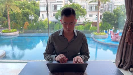 Asiatischer-Tausendjähriger-Unternehmer,-Der-Am-Laptop-Mit-Blick-Auf-Den-Luxuriösen-Pool-Arbeitet