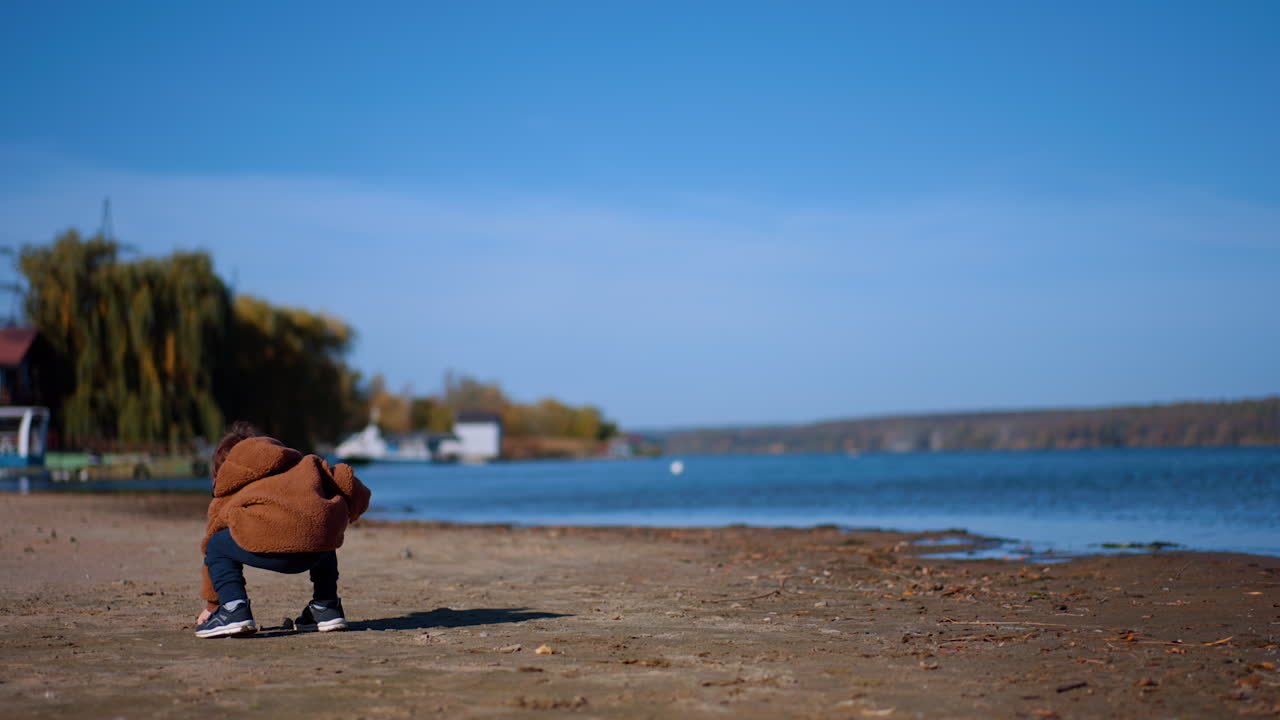 Rear view of a toddler clapping hands excitedly. Kid picks up a stone and brings it to the water.