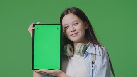 close up of asian teen girl student with a backpack smiling and showing green screen tablet to camera while standing in the green screen background studio