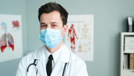 Portrait-Of-Handsome-Young-Man-Medic-In-White-Gown,-Medical-Mask-And-With-Stethoscope-Standing-In-Cabinet-And-Looking-At-Camera