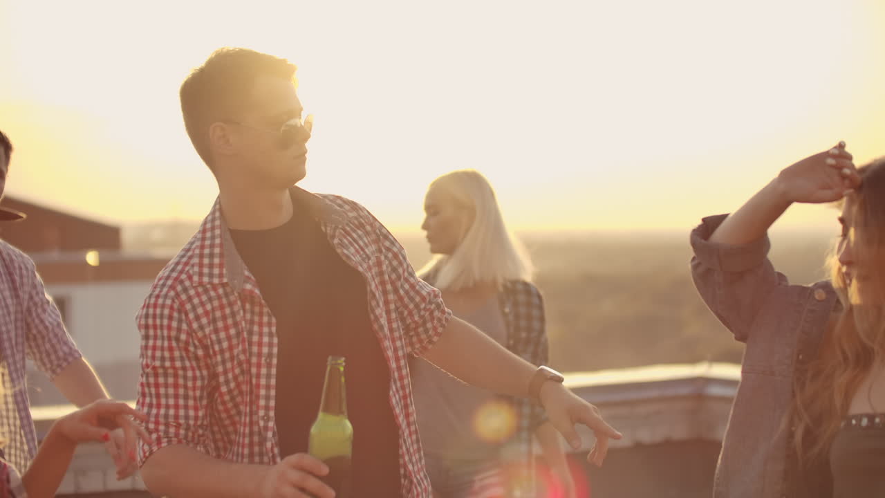 A young russian man in trendy glasses with beer moves in a dance on the party with his friends on the roof. He moves in dance with two girls.