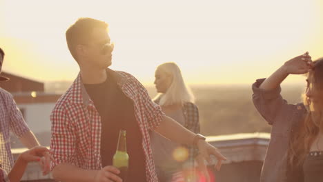 a young russian man in trendy glasses with beer moves in a dance on the party with his friends on the roof. he moves in dance with two girls.