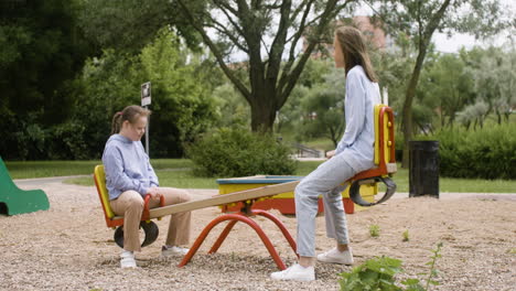 vista lateral de una niña con síndrome de down y su amiga rubia balanceándose en un mecedor de madera en el parque en un día ventoso