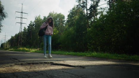 woman walking alone on quiet forest road under soft daylight, carrying black shoulder bag and wearing pink hoodie and jeans, surrounded by tall trees and power lines with visible roadside puddle