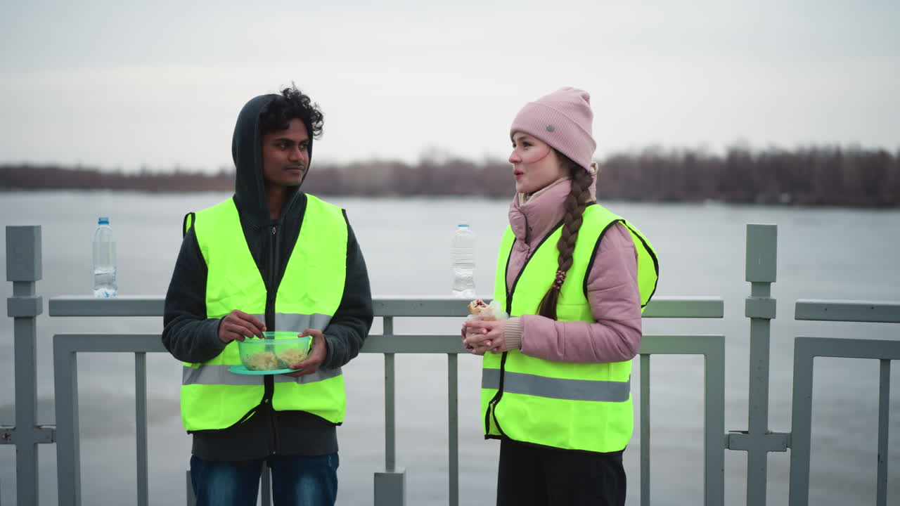 Man in hoodie and neon safety vest eating from container while woman in pink jacket and matching neon vest stands holding wrap on bridge near river during cold overcast day, representing outdoor work