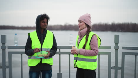 Man in hoodie and neon safety vest eating from container while woman in pink jacket and matching neon vest stands holding wrap on bridge near river during cold overcast day, representing outdoor work