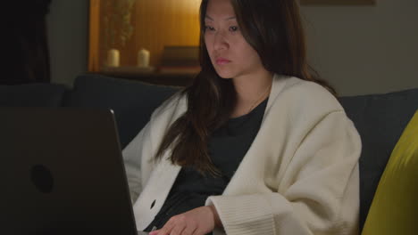 woman spending evening at home sitting on sofa with laptop computer looking at social media streaming or scrolling online