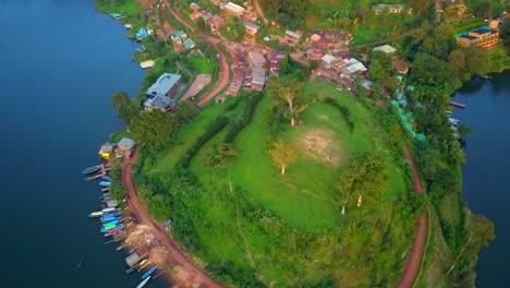 aerial top-down shot of a village on the northeastern shore of lake bunyonyi, uganda, showing traditional homes, vibrant green fields, and a colorful line of wooden boats at the lakeside dock