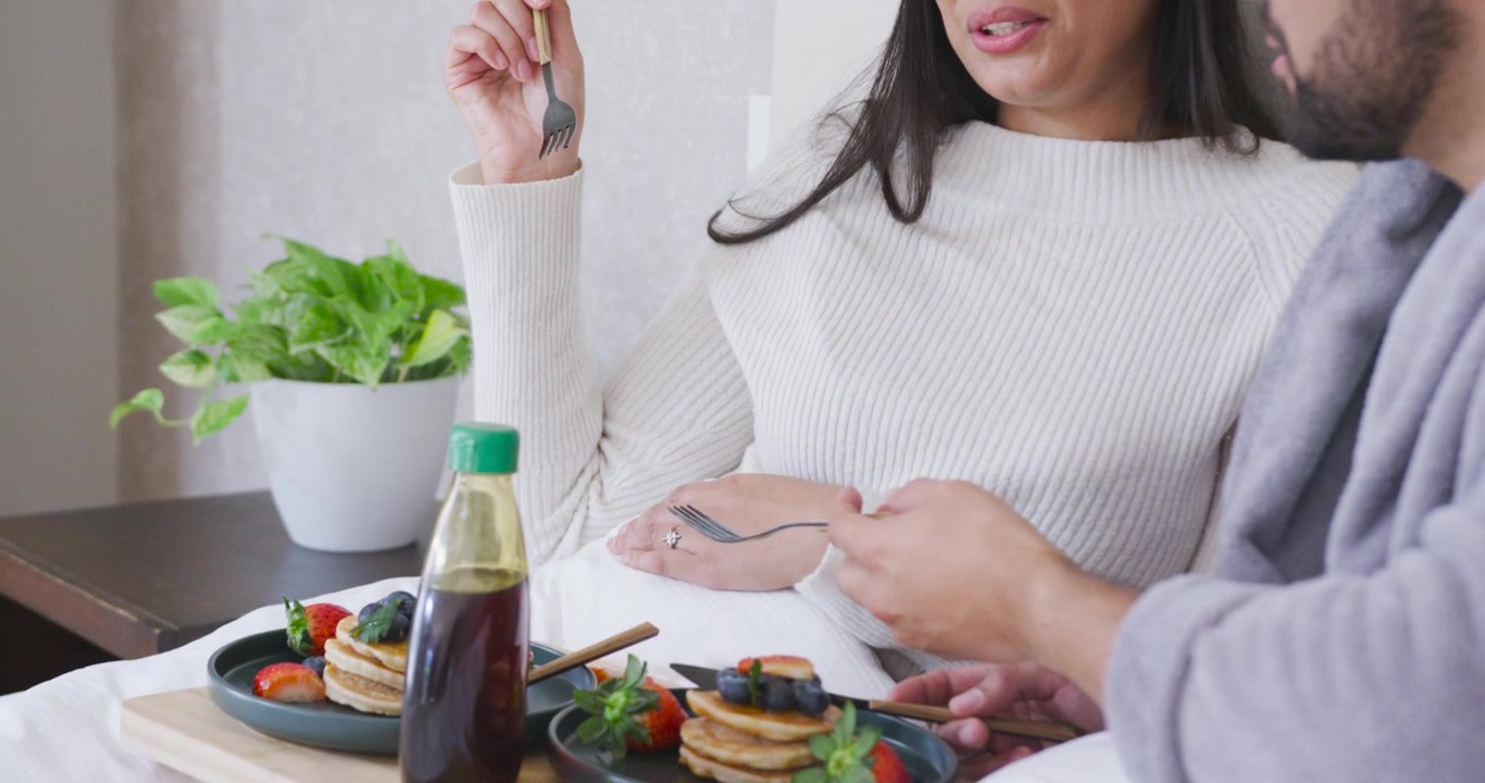 Coppia afroamericana a letto, la donna che alza una forchetta e parla, l'uomo che allunga la forchetta preparando i pancake.