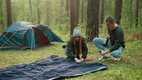 husband pumps tent bag as wife checks air level smiling while he looks at her warmly in peaceful forest camp setup with tent in background