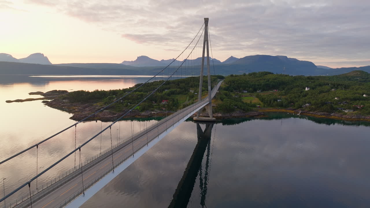 드론이 나르비크 다리를 오르고, 황금 시간 해가 지는 하늘이 평화로운 물에 반사됩니다.