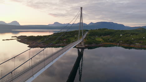 Drone-ascends-Narvik-bridge,-golden-hour-sunset-sky-reflected-on-peaceful-water