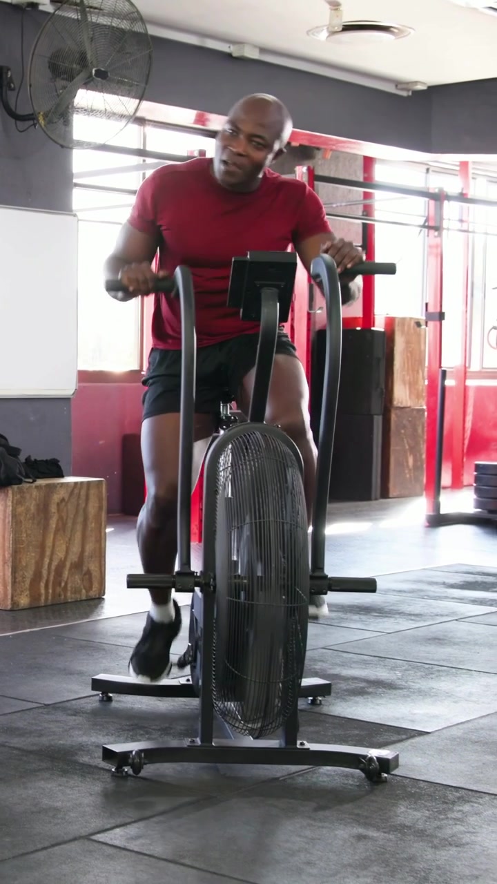 Video verticale: Uomo in maglietta rossa pedala con costanza, sforzandosi di più su una air bike in palestra per allenamento cardio.