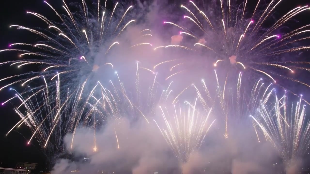Lancio di fuochi d'artificio come colpi di mortaio, che riempiono il cielo notturno del parco, con la folla che filma con i telefoni tra il fumo.