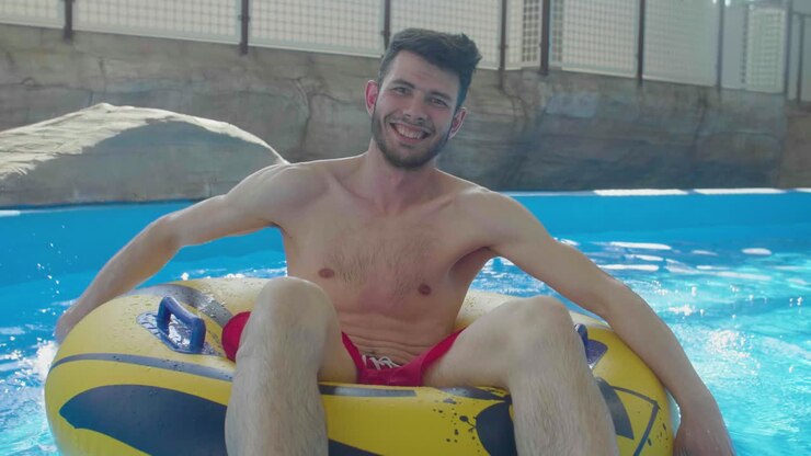 Young happy caucasian male sitting on inflatable lifebuoy ring rowing in swimming pool water slow motion portrait of funny summer holiday