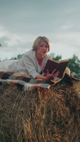 dama con la pierna levantada en una bala de heno con una manta a cuadros, leyendo un libro en el campo abierto con un hilo de heno en la boca, disfrutando de un momento pacífico al aire libre rodeado de naturaleza
