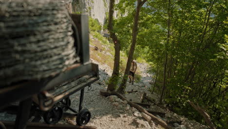 hiker walkig on a rocky path uphill in the shadow of the trees and continues to walk towards the little house like structures at the bottom of a sabotin clif