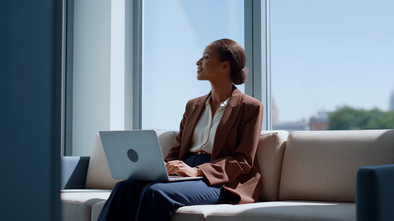 Woman Typing On Laptop By Sunlit Window, Relaxed Posture On Comfortable Sofa, Brown Blazer And Professional