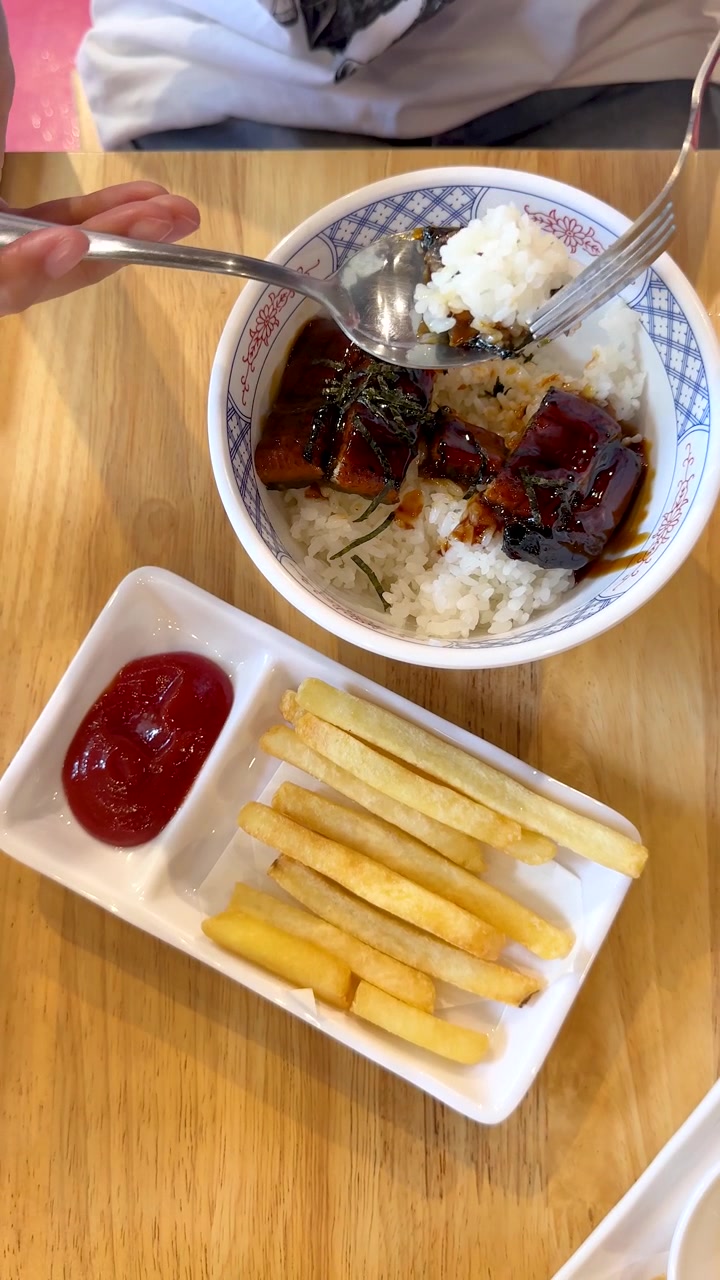 Seseorang makan mangkuk nasi belut bakar dengan kentang goreng dan saus tomat di restoran yang terang.