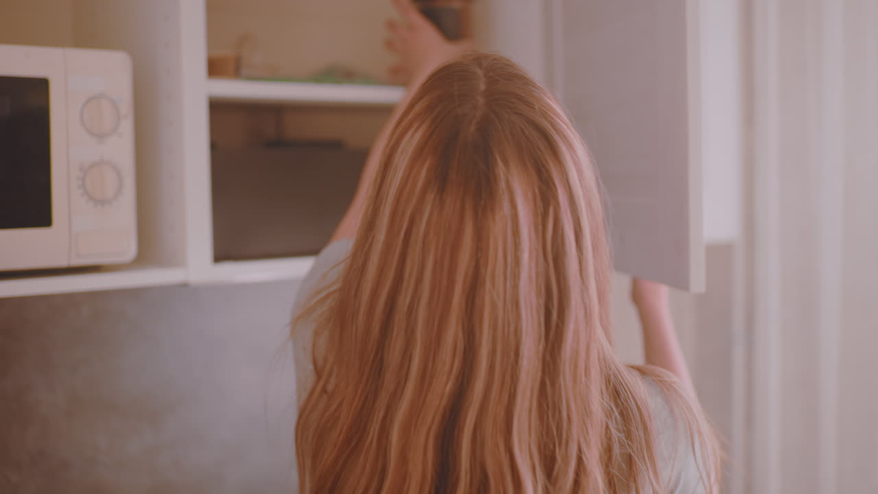 Natural light streams into kitchen as woman walks in carrying coffee bottle, places it on counter top and brings glass cup forward, creating warm serene morning atmosphere with soft glow indoors