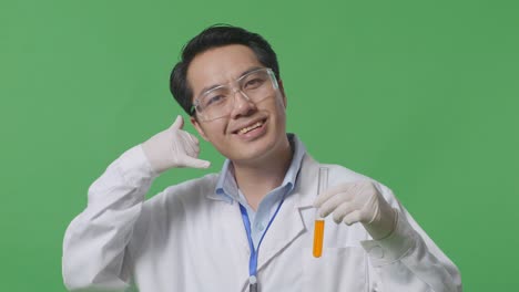 close up of asian man scientist with orange liquid in the test tube smiling and and making call me gesture while standing on the green screen background in the laboratory