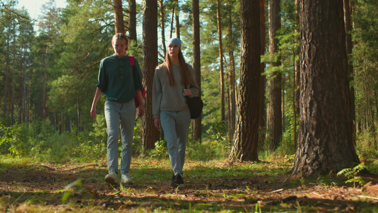 Friends walking through sunlit forest, one with red backpack and cloth, other with blue headscarf, focused expressions as sunlight filters through trees and reflects gently on their faces