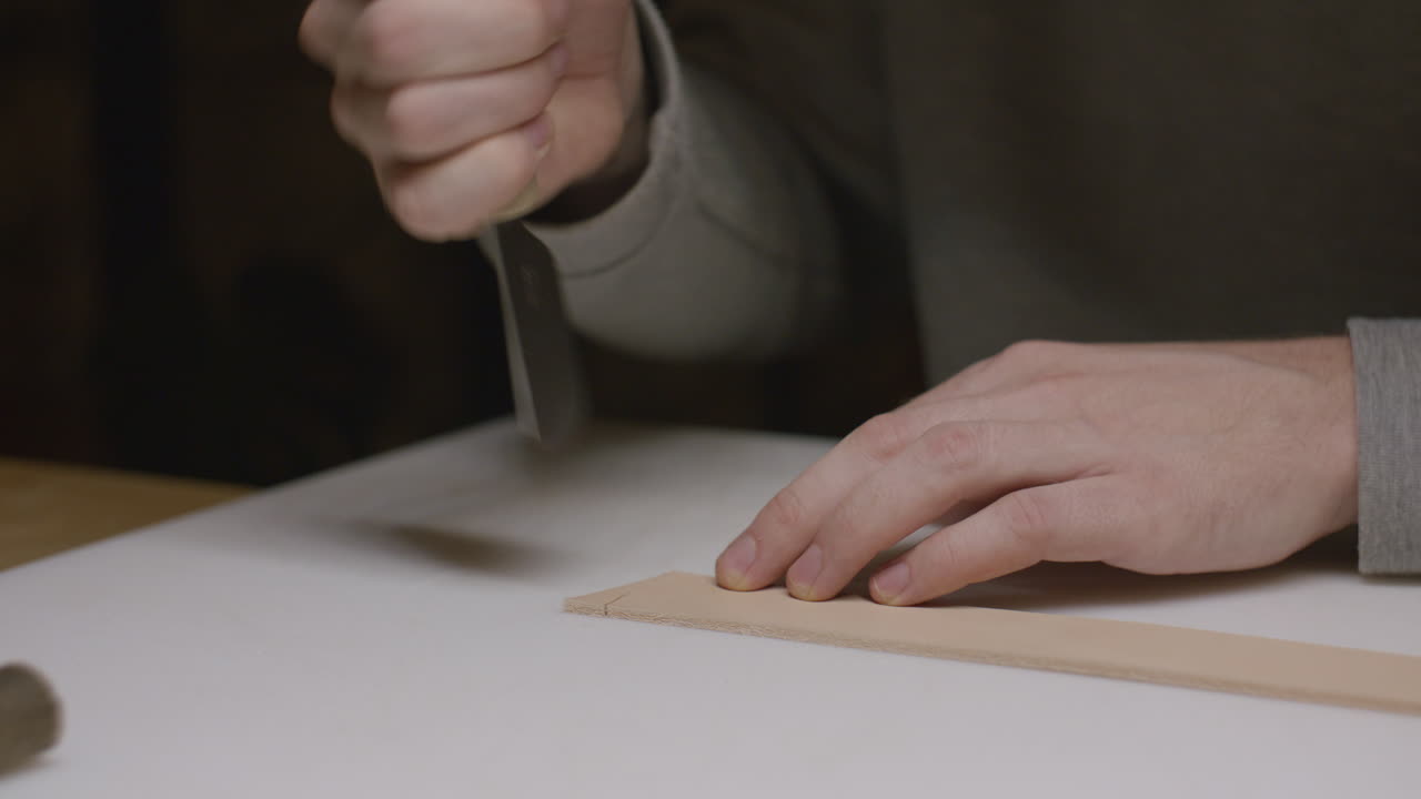 A craftsman carefully cuts a strip of leather using a sharp precision knife on a work surface, demonstrating skill and attention to detail in leather crafting.