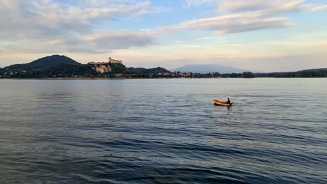 fisherman rowing small wooden fishing boat on maggiore lake in italy with angera fortress in background