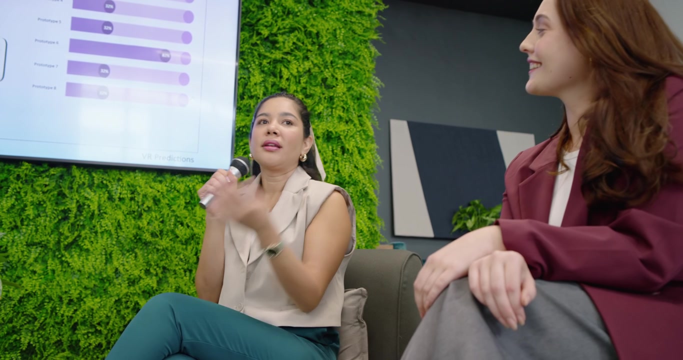 Panel di donne diverse che presenta con un tablet, posando mentre parla di VR, con una co-conduttrice che risponde al microfono in una lounge.
