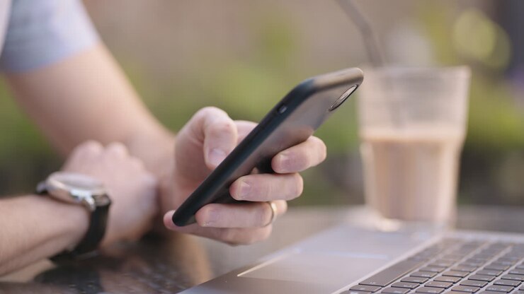 Primo piano di un uomo con un telefono cellulare che effettua operazioni bancarie online, fa acquisti su Internet e scrive messaggi sui social network, mentre è seduto con un portatile