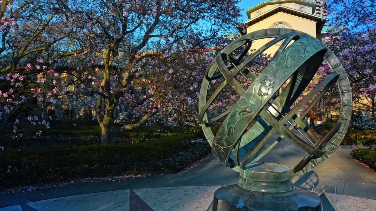 New York, USA, 8 aprile 2026: Primo piano di una sfera armillare in bronzo con sfondo di magnolie in fiore. Vista dettagliata di una meridiana a sfera armillare in bronzo con alberi di magnolia rosa a fuoco morbido
