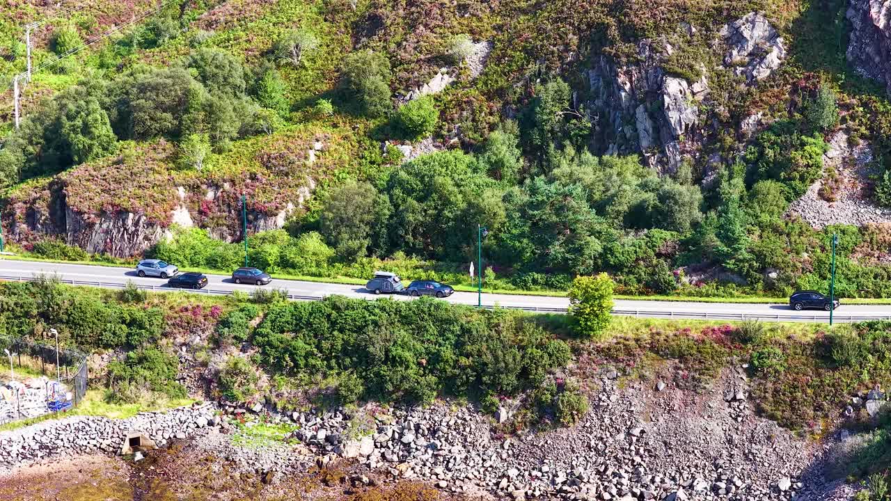 Multiple cars travel on a winding coastal road, lush greenery, rocky hillside, aerial daylight view