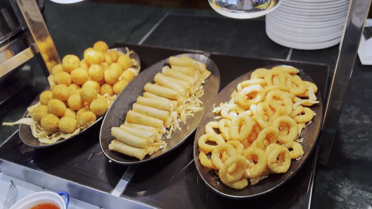 Close-up of golden-fried finger foods—cheese balls, rolls, and rings—on a buffet counter under warm lighting. Great for hospitality, food, or catering stock needs.