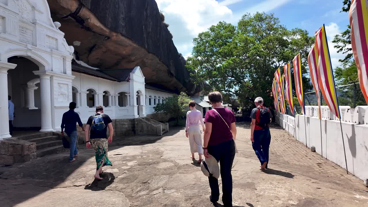 Visitors walk towards the historic Dambulla Cave Temple in Sri Lanka, adorned with colorful Buddhist flags fluttering in the breeze under a bright sky.