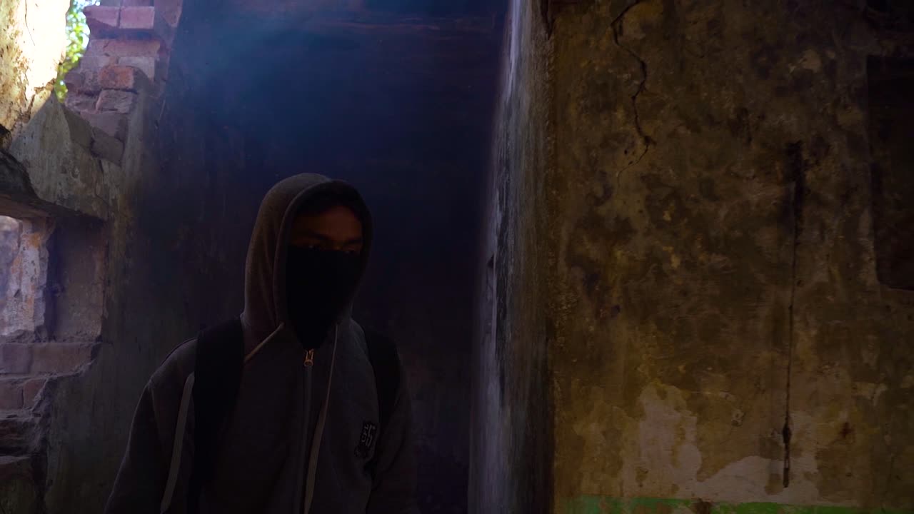 Vista frontale al rallentatore di un uomo coperto dalla testa ai piedi, che indossa una borsa e una maschera, mentre tiene una torcia per farsi strada lungo un sentiero buio e inquietante all'interno di un vecchio luogo.