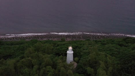 Aerial-drone-hovering-above,-capturing-hilltop-white-lighthouse-sending-off-beams-of-light-and-beautiful-townscape-and-coastal-seascape-in-the-evening-at-Xiaoliuqiu-Lambai-Island,-Pingtung,-Taiwan