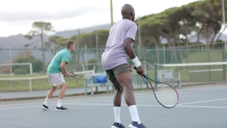 Two-diverse-male-friends-playing-doubles-returning-ball-on-outdoor-tennis-court-in-slow-motion