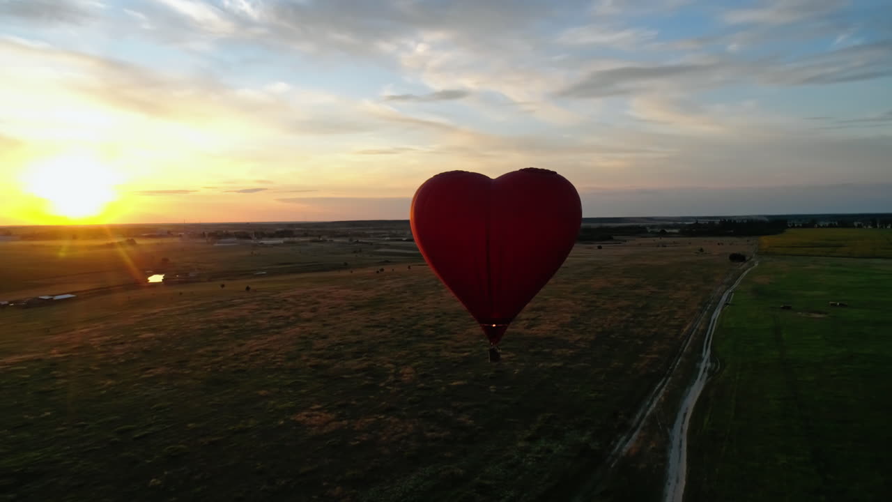 Heart shape aerostat against the setting sun. Red hot air balloon with basket flying over the green fields at sunset. Romantic airship in summer.