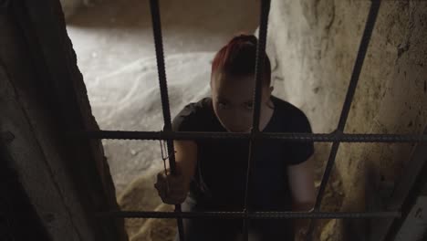 a young woman kneeling in a cell clutches the bars and raises her head to stare in a terrifying way