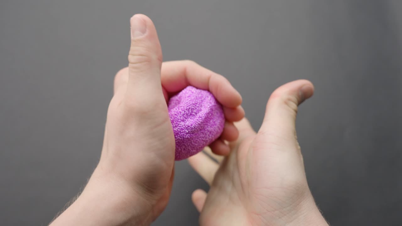 White hands knead purple floam slime, highlighting texture against a grey background.