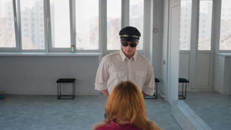 man in black cap and sunglasses wearing white shirt holds seated woman head while demonstrating gesture in front of large windows inside bright minimal studio room with reflective mirror background