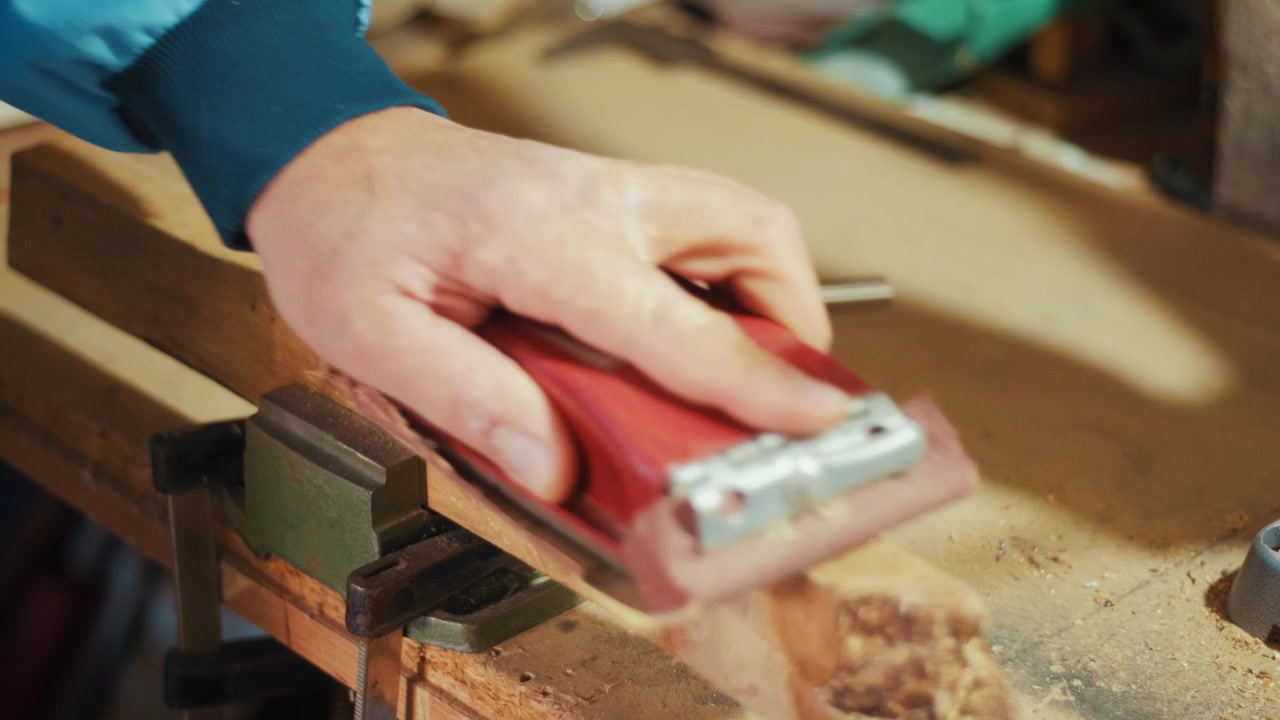 Close-up of a craftsman using a red sanding block to smooth a wooden plank by hand in a workshop. Authentic DIY woodworking footage for craft, carpentry and maker projects