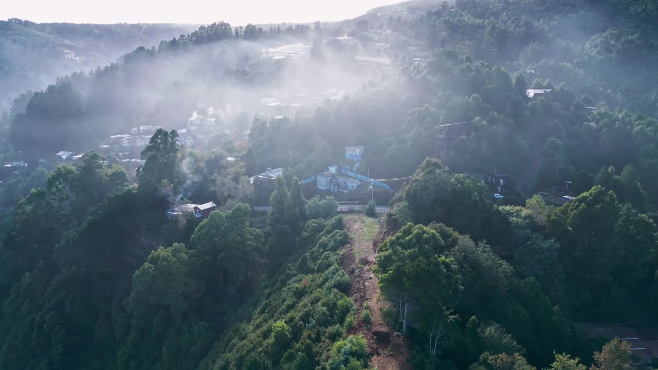 Şili kırsalında yemyeşil ormanlık tepelerde, atmosferik sabah sisiyle kaplı gizemli bir köyün drone ile geriye doğru çekilmiş havadan görüntüsü