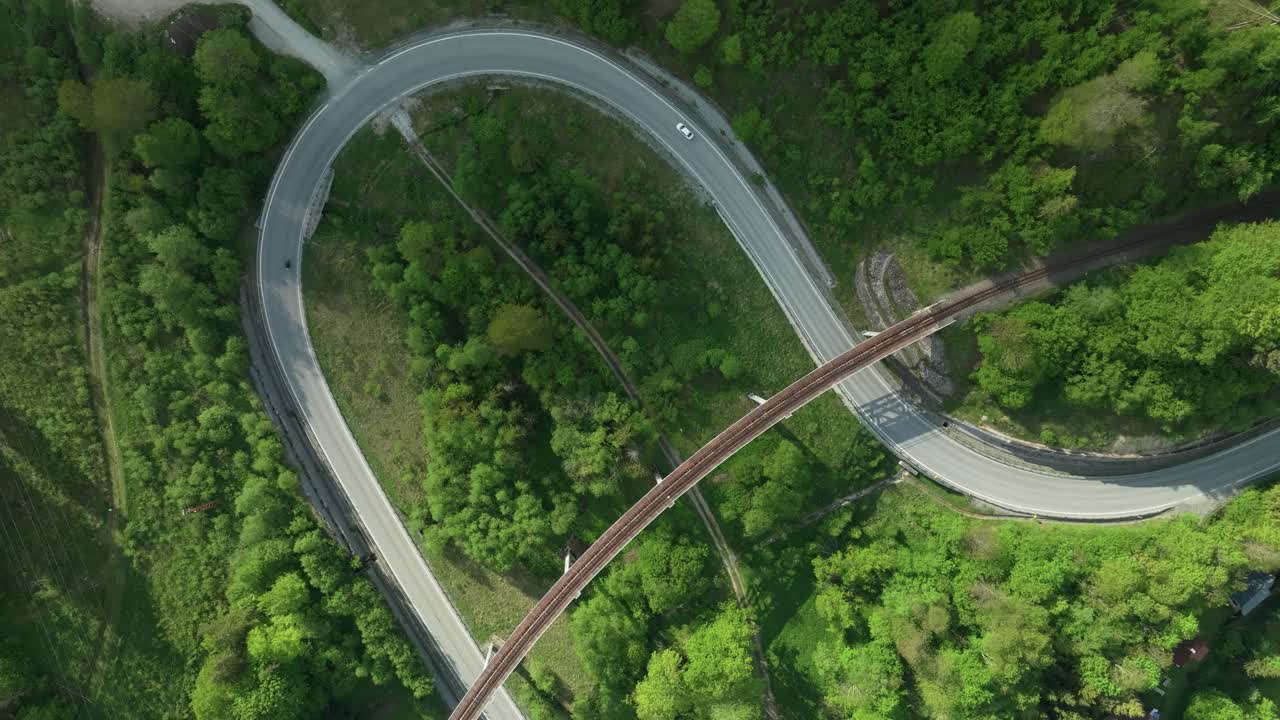An aerial overhead footage of a winding road intersected by an old railway bridge in the middle of a forest. Multiple cars are driving on a fairly empty road. Clouds create nice shadows. LuPa Creative