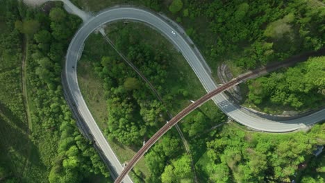 an aerial overhead footage of a winding road intersected by an old railway bridge in the middle of a forest. multiple cars are driving on a fairly empty road. clouds create nice shadows. lupa creative