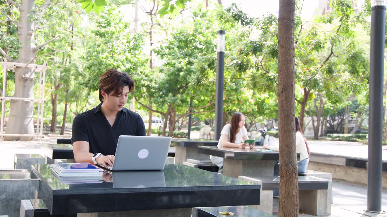 Seorang pria muda Thailand berbicara di teleponnya sambil bekerja dengan laptop di luar ruangan.