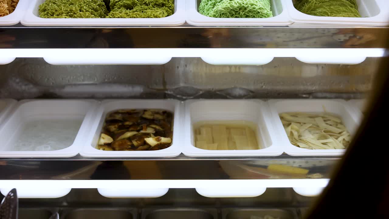 Person uses tongs to pick noodles and tofu from self-service buffet under bright lighting