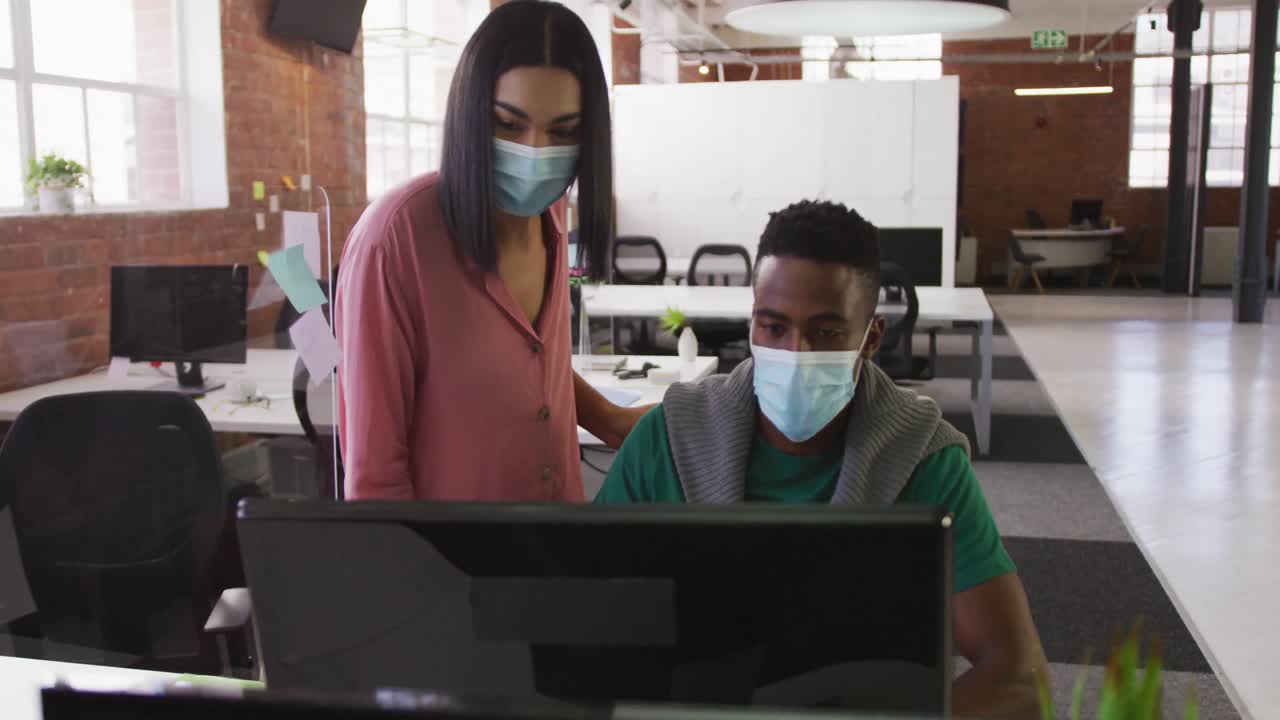 Diverse male and female business colleagues wearing face masks in discussion at work using computer