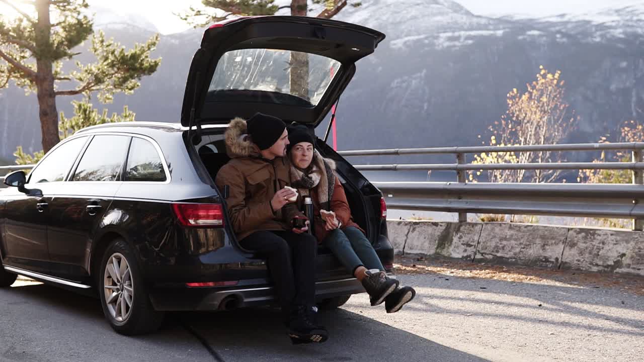 una pareja de enamorados en un viaje por carretera. el chico y la chica están sentados en el maletero abierto del coche, comiendo sándwiches. admirando la vista a su alrededor: la hermosa naturaleza de noruega. detenerse al aire libre en medio de la carretera
